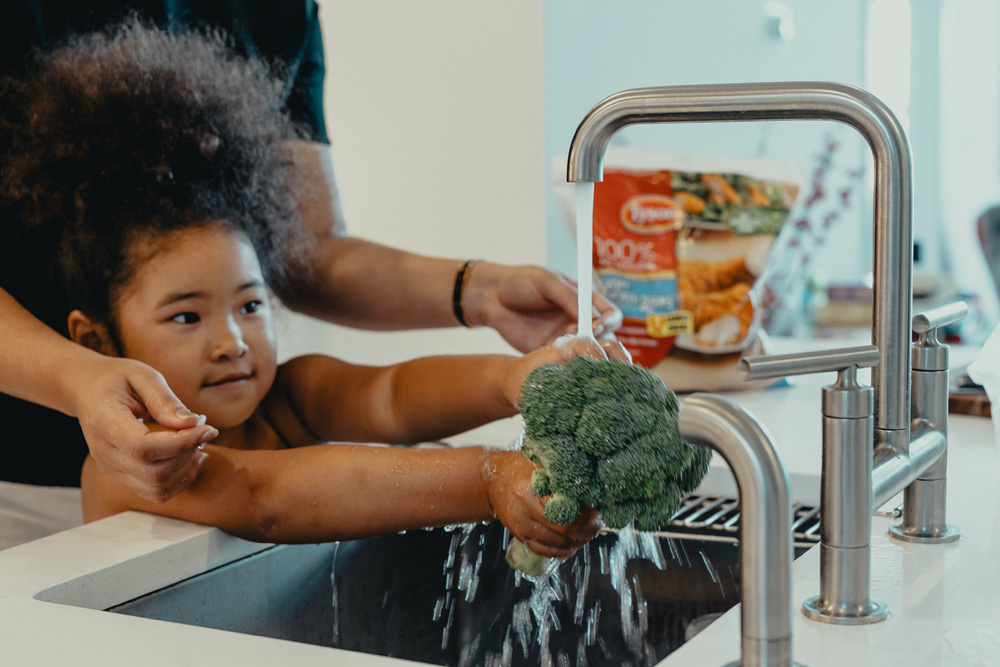 a girl washing broccoli under the kitchen sink while her mom helps